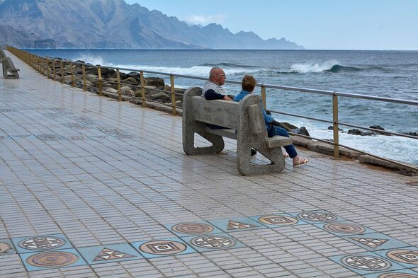 An older couple sits on a stone bench along a tiled coastal promenade, looking out at crashing ocean waves and dramatic mountain cliffs in the distanc