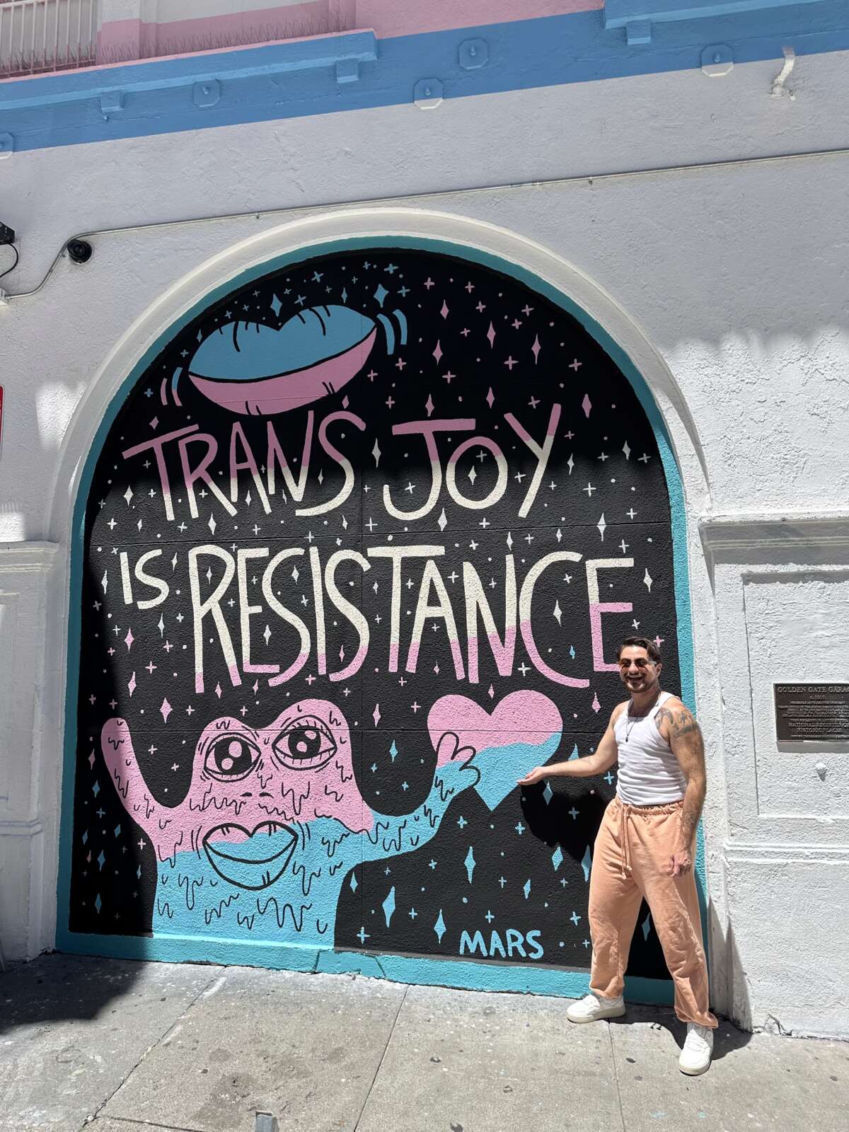 Trans artist and muralist, Mars Wright, standing in front of a mural he painted in San Francisco's Tenderloin Neighborhood in coordination with The Transgender District.