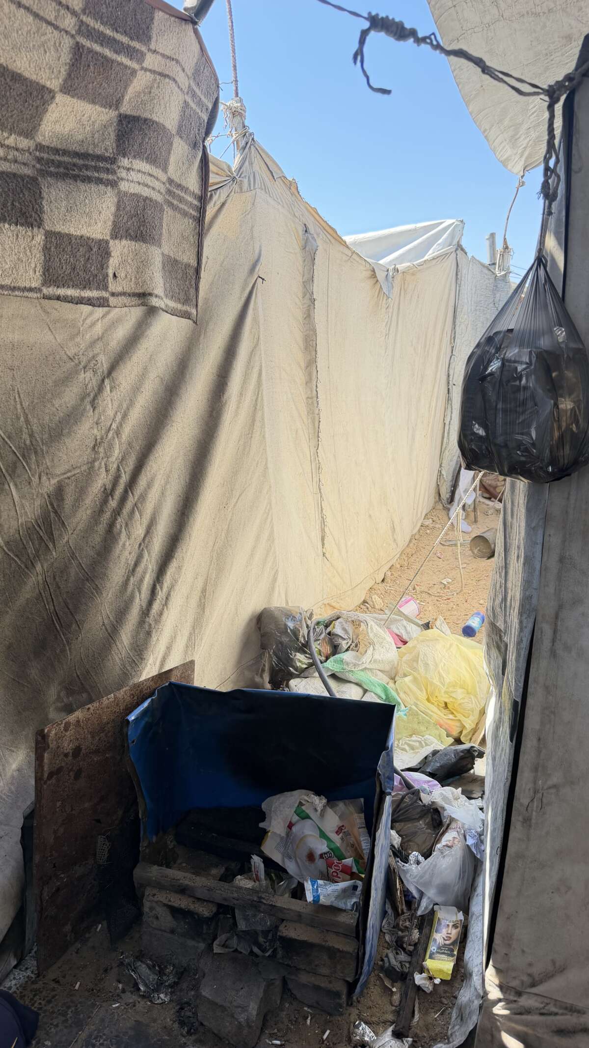 Khadra Al-Ghoul, 28, cooks over plastic food packaging scavenged from the street in a narrow alley between displaced families' tents in the Al-Masri camp northwest of Gaza City, where firewood is unaffordable and tight spaces pose constant fire risks.