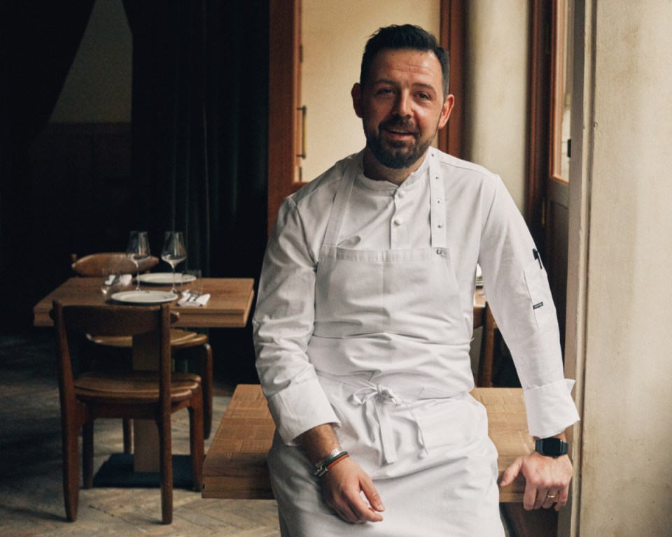 Smiling male chef in white uniform and apron, leaning on a wooden table in a restaurant dining room.