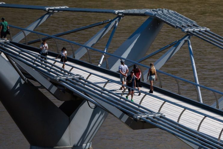 Pedestrians walk across a modern pedestrian bridge with steel cables and supports over brown water.