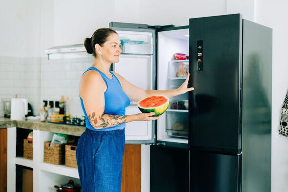 Woman holding watermelon by open refrigerator in kitchen