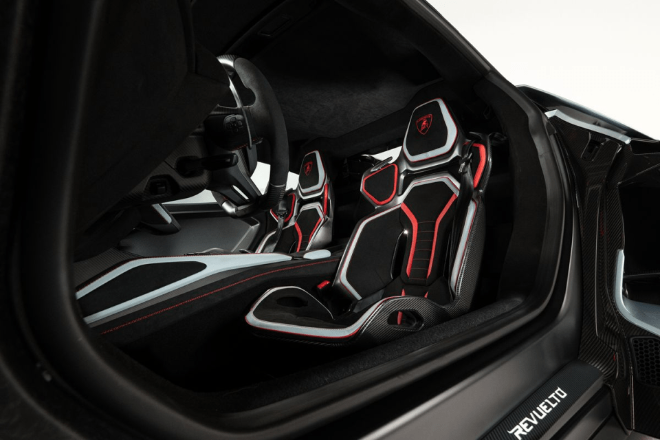Modern car interior with black and red racing seats, visible steering wheel, and REVUELTO on the sill.