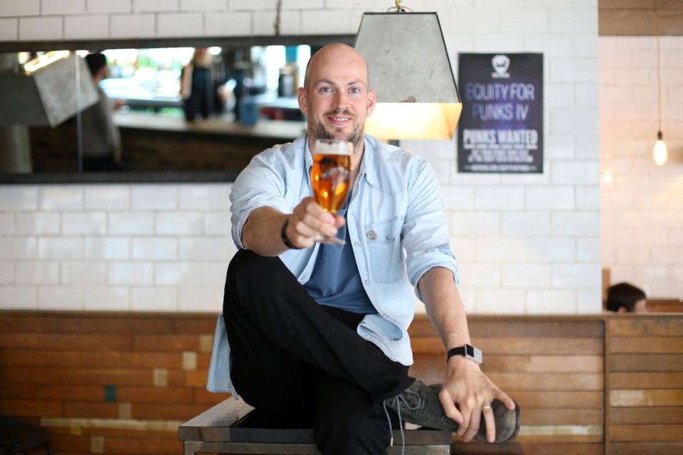 Bald man in blue shirt and black pants offering a glass of beer, smiling at the camera
