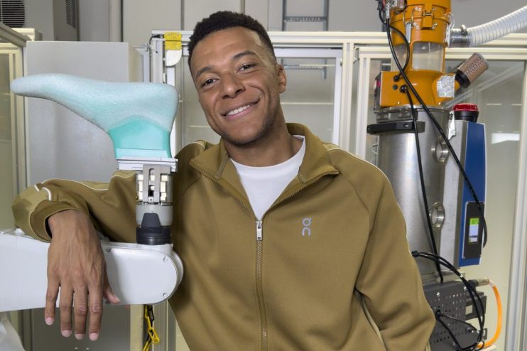 Smiling man in brown tracksuit leaning on a robotic arm with a 3D-printed shoe prototype in a lab