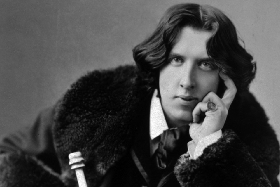 Black and white portrait of Oscar Wilde, a man with wavy hair, wearing a fur-collared coat and a ring, resting his chin on...
