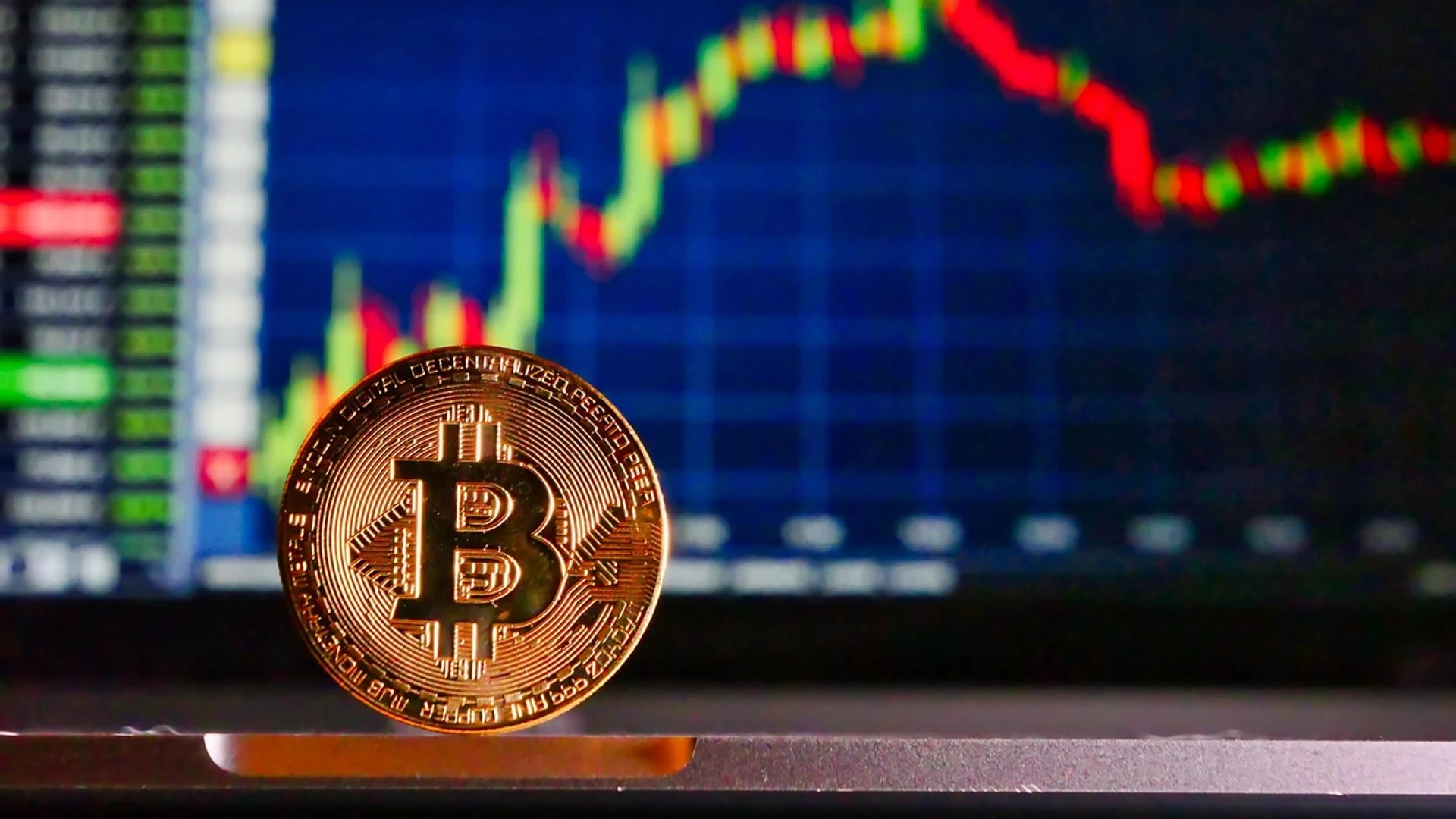 A physical Bitcoin coin in the foreground with a financial candlestick chart on a computer screen in the background.