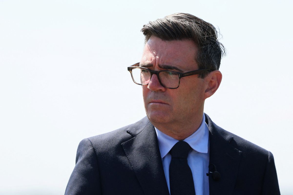 Andy Burnham, Mayor of Greater Manchester, in a suit and glasses, looking serious against a bright sky.
