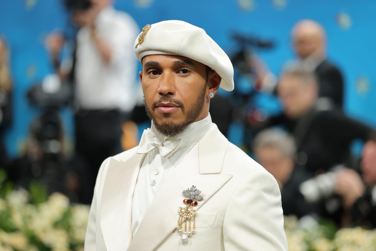 Lewis Hamilton in white suit, bow tie, beret, and pearl brooch looking directly at the camera.