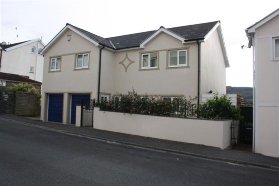 Modern cream-colored house in Wales with blue garage doors, white windows, and a landscaped front wall.